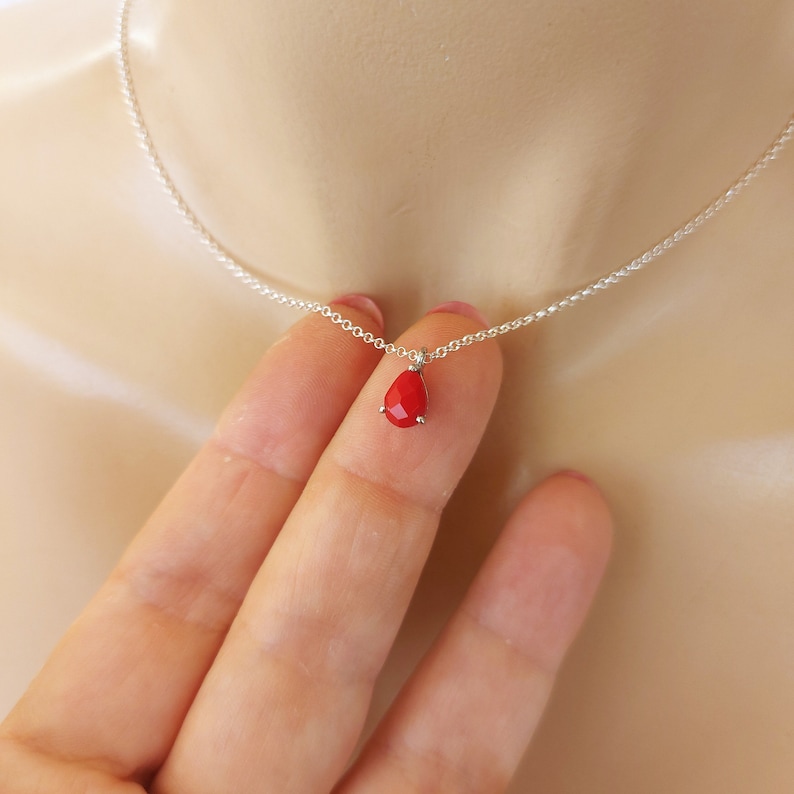 May include: A delicate silver chain necklace featuring a small, faceted, teardrop-shaped red pendant. The pendant is secured by a simple metal prong setting, suspended from a thin, polished silver-tone link chain that rests against the skin.