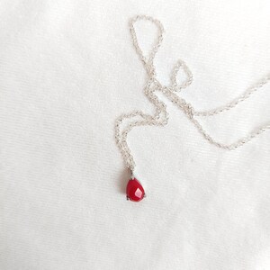 May include: A silver-toned chain necklace featuring a single, faceted, teardrop-shaped red pendant. The pendant is secured in a simple metal setting with small prongs, and the delicate chain is composed of small, interconnected circular links.