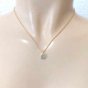 May include: A delicate gold chain necklace featuring a small, teardrop-shaped light blue gemstone pendant. The pendant is secured in a simple gold setting with three prongs, suspended from a thin, polished gold-tone metal link chain.
