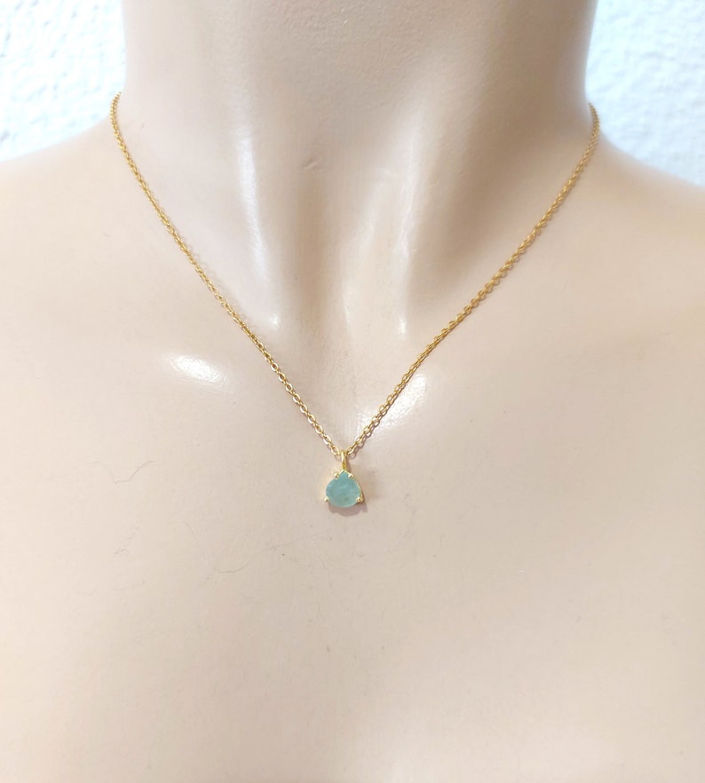 May include: A delicate gold chain necklace featuring a small, teardrop-shaped light blue gemstone pendant. The pendant is secured in a simple gold setting with three prongs, suspended from a thin, polished gold-tone metal link chain.