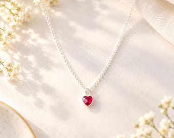 May include: A delicate silver chain necklace featuring a small, faceted heart-shaped red gemstone pendant. The necklace is crafted with a fine link chain and a polished metal setting that secures the vibrant red heart charm at the center.