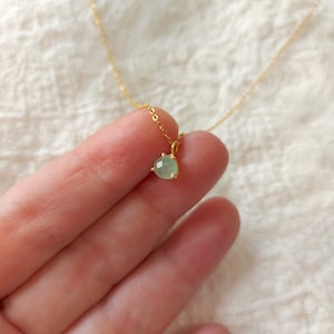 May include: A delicate gold chain necklace featuring a small, round, faceted light green gemstone pendant held in a simple gold prong setting. The pendant is suspended from a thin, polished gold-tone metal chain that extends upwards.