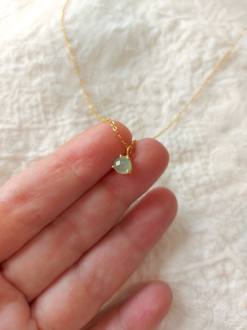 May include: A delicate gold chain necklace featuring a small, round, faceted light green gemstone pendant held in a simple gold prong setting. The pendant is suspended from a thin, polished gold-tone metal chain that extends upwards.