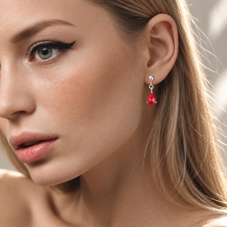 May include: A silver stud earring featuring a small, faceted, teardrop-shaped red stone that hangs from a polished silver ball post. The earring is worn in the earlobe, showcasing the vibrant red color and the reflective quality of the stone.