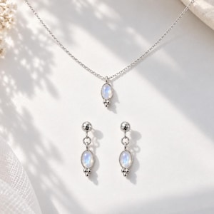 Pode incluir: Um conjunto de joias em prata composto por um colar de corrente fina e um par de brincos pendentes a condizer. Cada peça apresenta um pequeno cabochão de pedra da lua oval e iridescente com três pequenas esferas de prata na base.