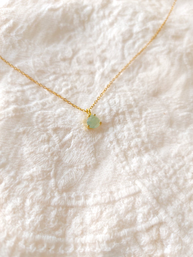 May include: A delicate gold chain necklace featuring a single, small, teardrop-shaped light green gemstone pendant held in a gold prong setting. The necklace is presented as a simple, refined piece of jewelry with a thin, polished gold metal chain.
