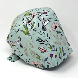 May include: A light blue scrub cap featuring a floral and hummingbird pattern. The design includes pink flowers, green leaves, and detailed illustrations of hummingbirds. The cap is made of a soft fabric and has a tie in the back.