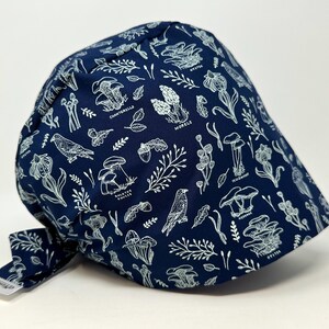 Könnte beinhalten: Marineblaue Kappe mit weißem botanischem Muster mit Pilzen, Blättern und Vögeln. Die Kappe hat einen Bindeband am Rücken und ein kleines Etikett an der Seite. Das Design enthält die Wörter "Chanterelle" und "Morels".