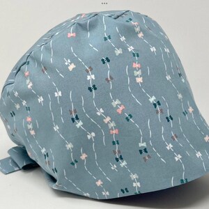 May include: A light blue scrub cap with a repeating pattern of bows in pink, gray, and teal, connected by white dashed lines. The cap has a rounded shape and a tie in the back.