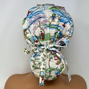 May include: A patterned surgical scrub cap featuring a Japanese landscape design with figures, trees, and water elements in shades of blue, green, red, and beige. The cap has a tie closure at the back.