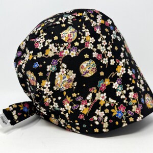 May include: Black surgical scrub cap with a colorful floral pattern. The cap features a repeating design of white, yellow, pink, and blue flowers and decorative elements on a black background. The cap has a tie in the back.