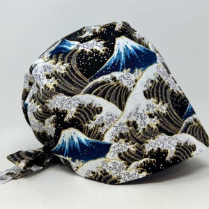 May include: A scrub cap featuring a Japanese-inspired print of stylised dark blue waves with white foam and mountain peaks against a black background. The fabric has a textured appearance and includes a small white tag on the side tie.