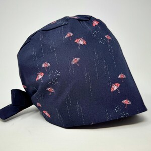May include: A navy blue surgical scrub hat with a tie, featuring a repeating pattern of pink and white umbrellas with white raindrops.