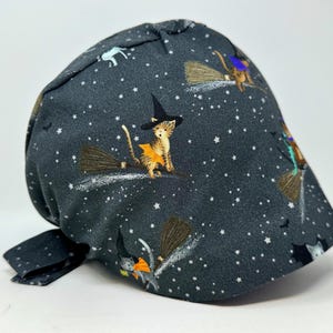 May include: Dark gray Halloween themed scrub cap featuring a pattern of flying witch cats with brooms, stars, and bats. The cats wear witch hats and orange capes. The cap has a tie in the back.