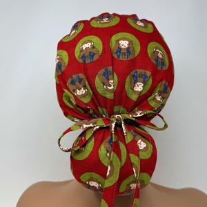 May include: A red scrub cap with a repeating pattern of monkeys in green circles. The cap is tied in a bow at the back. The monkeys are brown and wearing blue outfits.