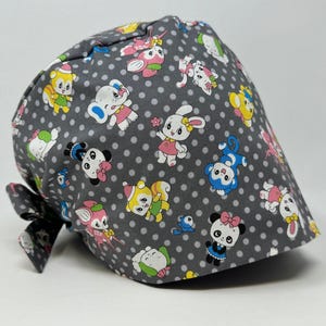 May include: A grey scrub cap featuring a repeating pattern of cartoon animals and white polka dots. The animals include bunnies, pandas, elephants, and squirrels in various colours. A black bow is attached to the back of the cap.