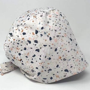 May include: A white patterned cap with a terrazzo design. The cap features a repeating pattern of various shapes in shades of black, gray, peach, and brown. The cap has a tie closure.