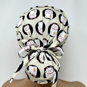 May include: A beige scrub cap featuring a repeating pattern of white and red Maneki-Neko cats within black circles. The cap is tied at the back with a matching bow. The cat design is a symbol of good luck and fortune.