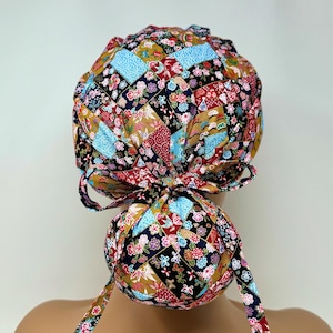 Könnte beinhalten: Eine bunte Patchwork-OP-Haube mit floralem und geometrischem Muster. Die Haube weist eine Vielzahl von Farben auf, darunter Blau, Rot, Gold und Schwarz. Die Haube ist hinten mit einer Schleife gebunden und hat lange Bänder.