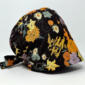 May include: A black scrub cap with a floral pattern. The design includes yellow, orange, and white flowers, with green and purple foliage. The cap has a tie closure. The fabric is soft.