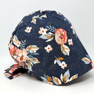 May include: A navy blue cap with a floral pattern featuring peach, white, and blue flowers. The cap has a brim and a tie in the back. The fabric has small gold dots.