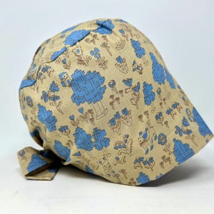 May include: A beige fabric hat with a repeating pattern of blue pixelated trees, flowers, and animal figures. The hat has a rounded crown and a small brim. The fabric is soft and appears to be cotton or a similar material.