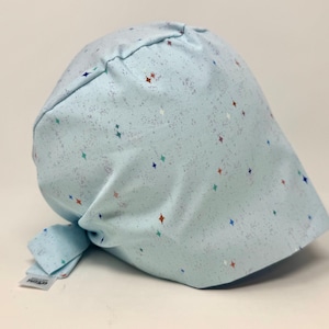 May include: A light blue scrub cap featuring a scattered pattern of small, multi-colored four-pointed stars and subtle grey speckles. The cap has a rounded crown, a wide front band, and fabric ties at the back for an adjustable fit, with a small white tag.
