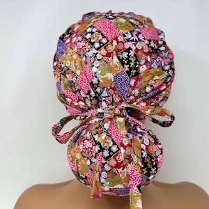 Könnte beinhalten: Eine bunte OP-Haube mit Patchwork-Design. Die Haube zeigt eine Vielzahl von Blumen- und geometrischen Mustern in Rosa, Gold, Schwarz und Lila. Sie hat einen Bindebandverschluss für eine verstellbare Passform.