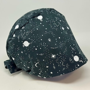 May include: A dark charcoal gray scrub cap featuring a white celestial print pattern of stars, planets, crescent moons, and constellations. The head covering has a rounded crown with stitched seams and a tie-back closure at the nape of the neck.