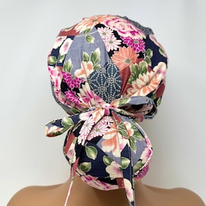 Könnte beinhalten: Eine OP-Haube mit floralem Muster, das eine Mischung aus rosa, orangefarbenen und weißen Blumen auf marineblauem Hintergrund zeigt. Die Haube hat eine Schleife am Rücken und Bindebänder. Das Design umfasst grüne Blätter und geometrische Formen.
