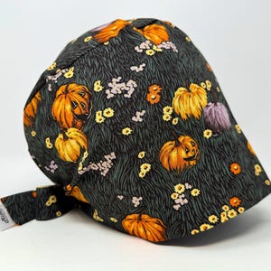 May include: A dark green cap featuring a Halloween design with orange pumpkins, yellow and purple flowers, and green grass. The cap has a curved brim and a small tag.