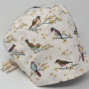 May include: A white fabric cap with a repeating pattern of birds and yellow flowers. The birds are in shades of brown, red, and blue. The cap has a tie closure. The design is reminiscent of a botanical illustration.