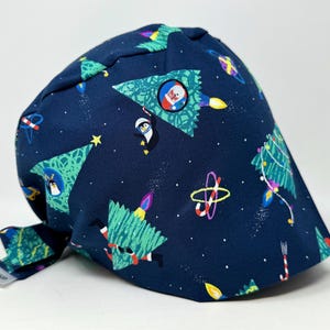 May include: A navy blue scrub cap with a whimsical Christmas-themed pattern. The design features green Christmas trees, penguins, rockets, and other festive elements. The cap is made of a soft fabric and has a tie in the back.