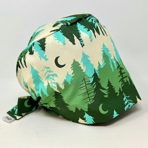May include: A forest-themed scrub cap featuring a repeating pattern of pine trees in shades of green and teal against a cream background. The cap includes a crescent moon design and a tie closure. The fabric is soft and flexible.