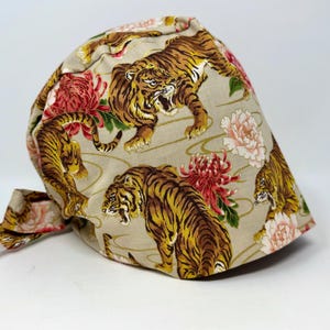 May include: A beige fabric cap featuring a repeating pattern of tigers, red and pink flowers, and green leaves. The tigers are brown and yellow with black stripes. The flowers are in full bloom. The cap has a tie in the back.