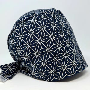 Könnte beinhalten: Eine marineblaue OP-Haube mit einem weißen geometrischen Sternmuster. Die Haube wird hinten mit einem passenden Stoffband gebunden. Ein kleines Etikett ist am Band befestigt.