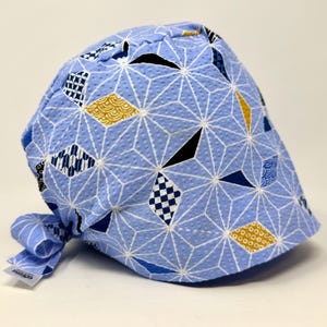 May include: A light blue scrub cap featuring a white geometric star pattern overlaid with scattered diamond shapes in navy blue, white, and mustard yellow. The fabric has a textured finish, and the cap includes a tie-back closure with a small white tag.