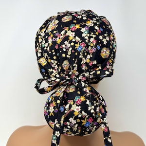 Peut inclure: Un bonnet de chirurgien noir avec un motif floral composé de fleurs blanches, jaunes, roses et bleues. Le bonnet est noué par un nœud à l'arrière. Le tissu semble être du coton.