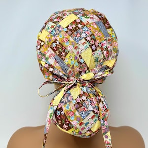 Könnte beinhalten: Eine bunte OP-Haube mit Patchwork-Design. Die Haube weist eine Vielzahl von floralen und geometrischen Mustern in Gelb-, Braun-, Rot- und Blautönen auf. Die Haube wird hinten mit passenden Stoffbändern gebunden.