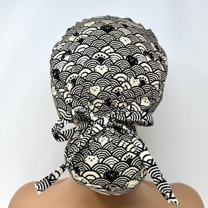 May include: A black and white patterned bouffant surgical scrub cap. The cap features a repeating wave pattern with cat faces. The cap is tied in the back with two long ties. The background is white.