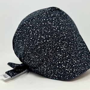 May include: A black scrub cap featuring a white speckled pattern resembling stars. The cap has a rounded crown and a tie-back closure at the nape. A small white fabric tag with the text "Mina" is visible on the side near the tie.