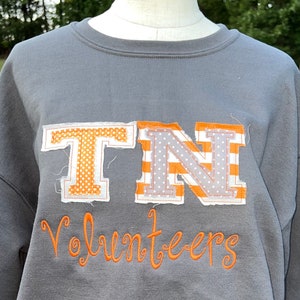 May include: Grey sweatshirt with a large orange and white "TN" appliqu&eacute; and the word "Volunteers" embroidered in orange below.