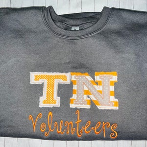 May include: A grey sweatshirt with the letters "TN" in orange and white fabric, with the word "Volunteers" embroidered in orange below.