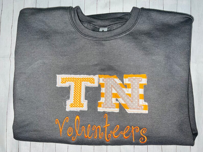 May include: A grey sweatshirt with the letters "TN" in orange and white fabric, with the word "Volunteers" embroidered in orange below.