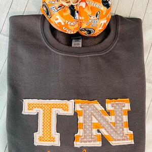 May include: A grey sweatshirt with a large orange and white "TN" appliqu&eacute; and the word "Volunteers" embroidered in orange below. The sweatshirt is decorated with a small orange and white pumpkin with a houndstooth pattern.