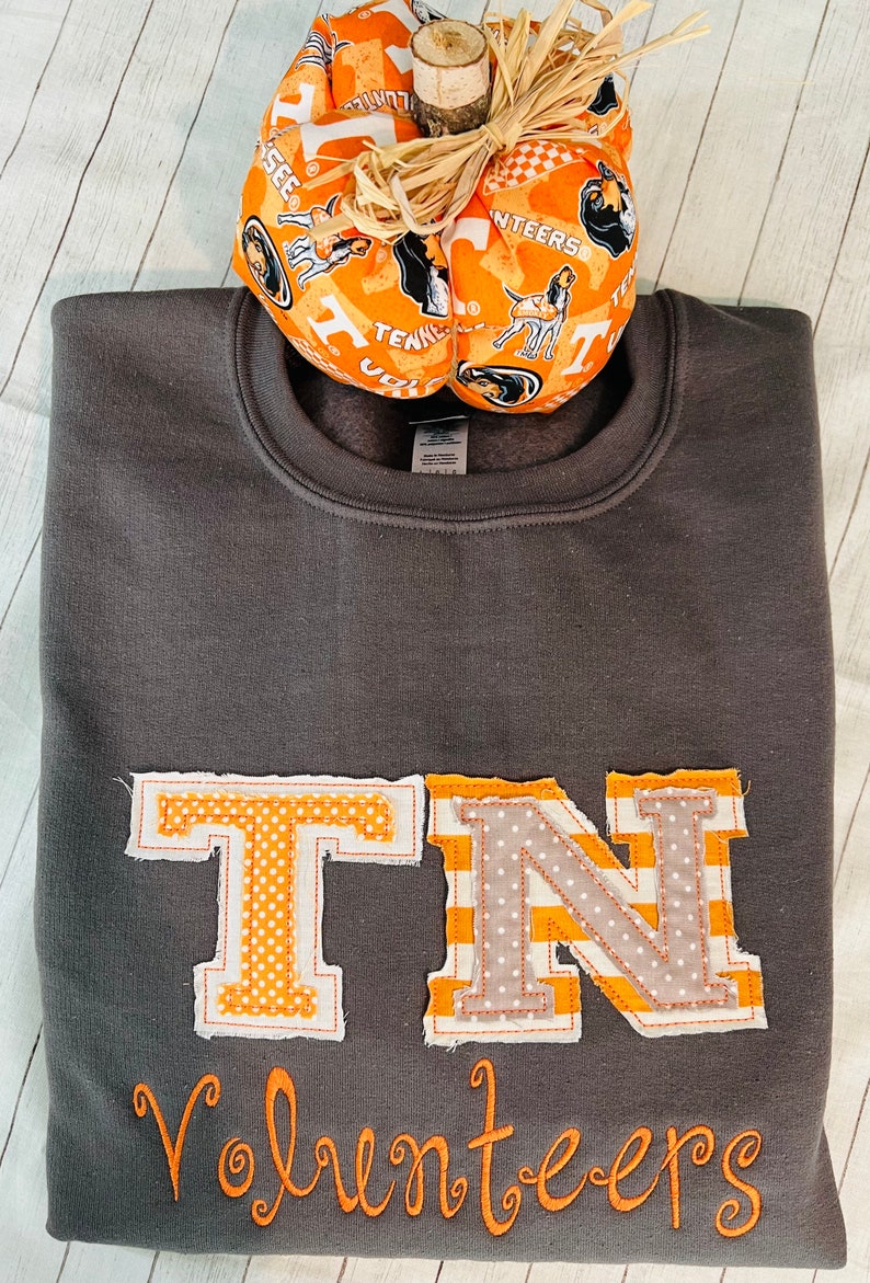 May include: A grey sweatshirt with a large orange and white "TN" appliqu&eacute; and the word "Volunteers" embroidered in orange below. The sweatshirt is decorated with a small orange and white pumpkin with a houndstooth pattern.