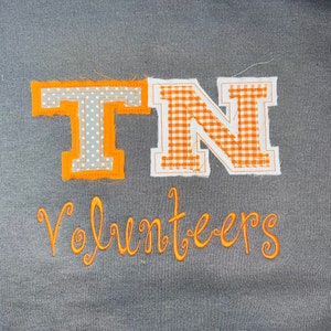 May include: Orange and white fabric letters spelling "TN" against a grey background. The letters are stitched onto the fabric and have a gingham pattern. Below the letters is the word "Volunteers" in orange thread.