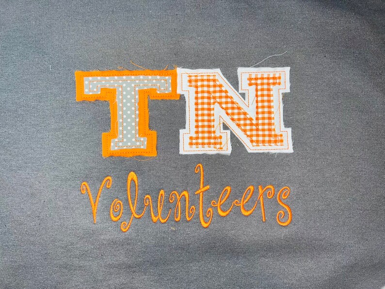 May include: Orange and white fabric letters spelling "TN" against a grey background. The letters are stitched onto the fabric and have a gingham pattern. Below the letters is the word "Volunteers" in orange thread.