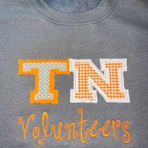 May include: A grey sweatshirt with an orange embroidered design. The design features the letters "TN" in a gingham pattern and the word "Volunteers" in a script font.