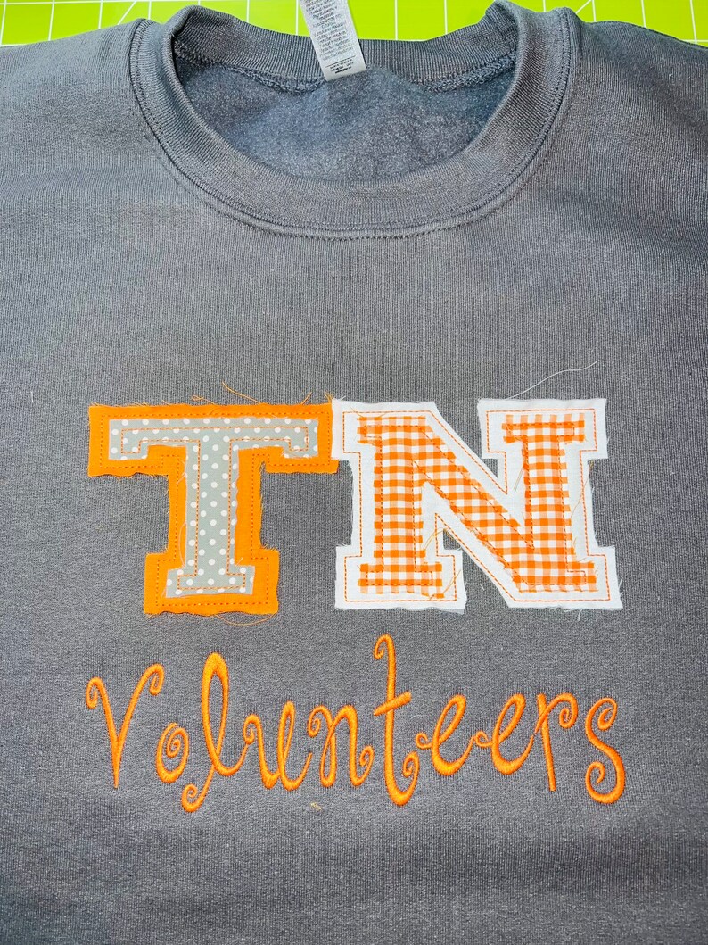 May include: A grey sweatshirt with an orange embroidered design. The design features the letters "TN" in a gingham pattern and the word "Volunteers" in a script font.